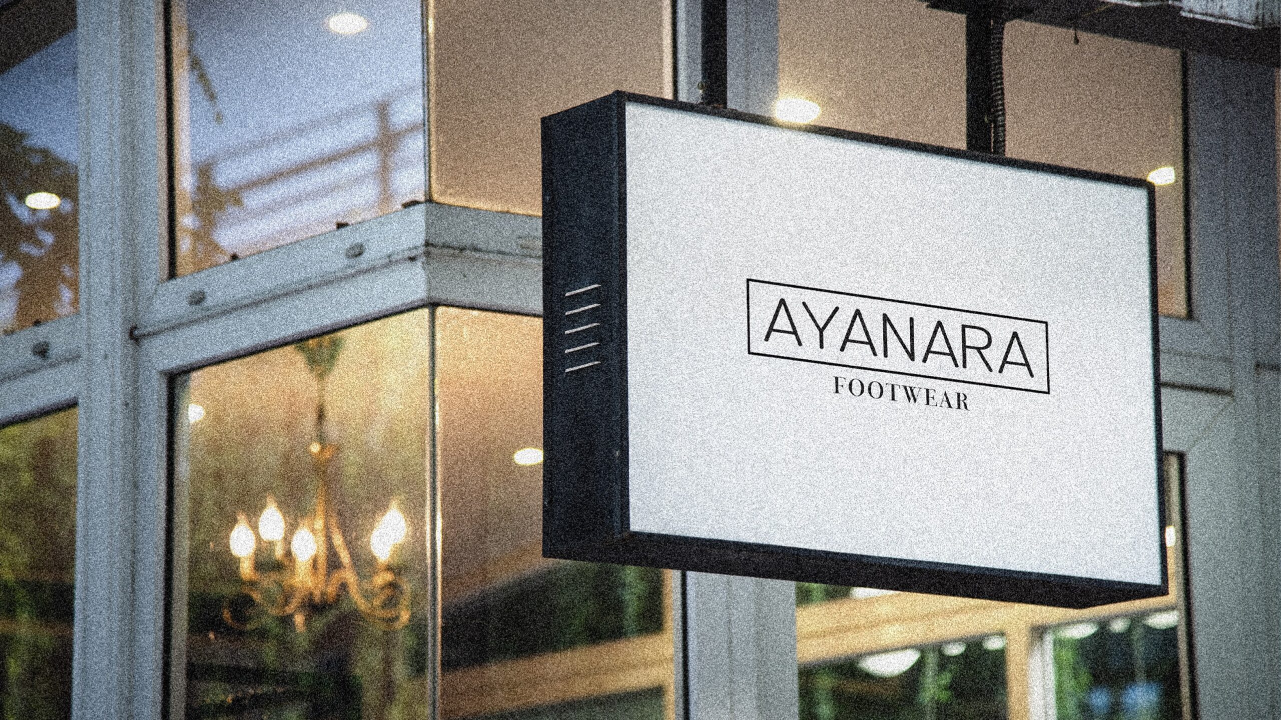 An outdoor illuminated signage for "AYANARA FOOTWEAR" displayed on a modern storefront. The design features a clean, minimalist black-and-white logo, conveying elegance and simplicity. The sign is mounted against a glass window, reflecting warm indoor lighting, including a classic chandelier, adding a cozy and inviting atmosphere. The image captures a blend of sophistication and contemporary retail branding.