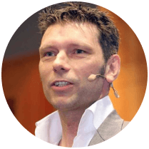 A close-up image of a man wearing a suit and a headset microphone, speaking confidently in a professional setting.