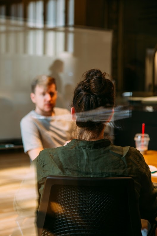 Two people having a conversation in a modern office setting, with a glass panel creating a blurred reflection effect, and a red straw cup visible on the table.