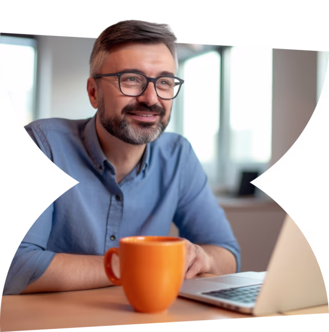 Smiling man wearing glasses, sitting at a desk with a laptop and orange coffee mug in a bright office setting