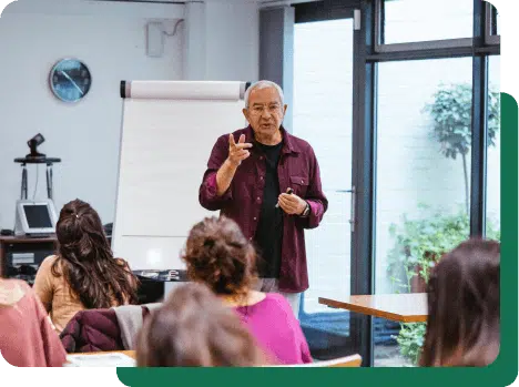 Instructor giving a lecture to a group of students in a classroom setting with large windows and a flip chart