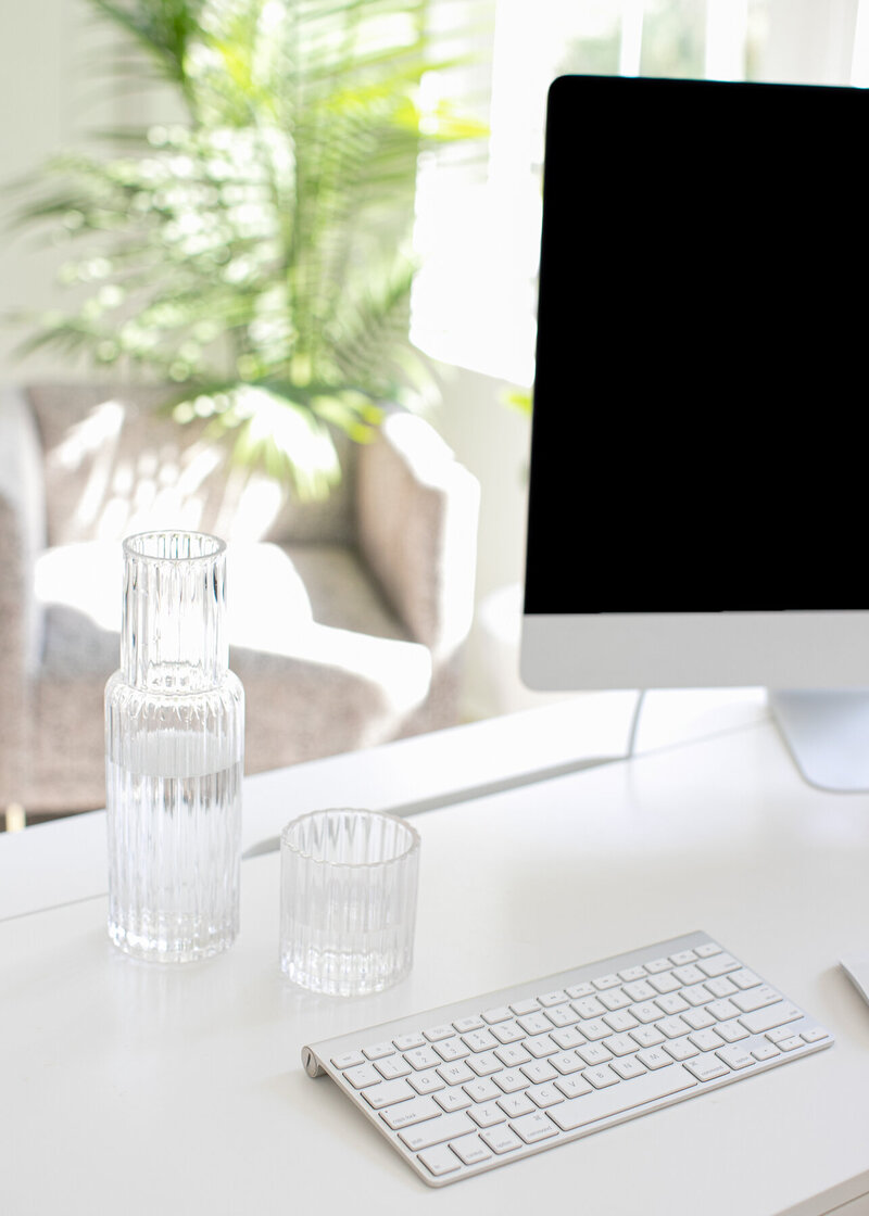 Minimalist workspace with a computer, keyboard, water pitcher, and glass on a white desk.