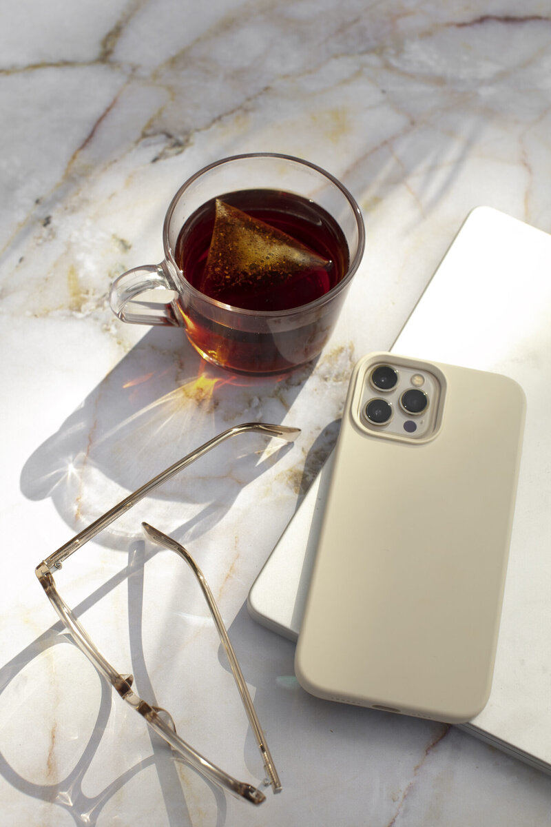 iPhone 14 Pro in a beige case, eyeglasses, and a cup of tea on a marble table.